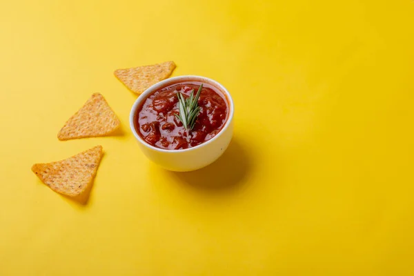 Red sauce with rosemary served in bowl with nacho chips on yellow background with copy space. unaltered, unhealthy food, snack, dipping sauce, herb, crunchy and savory food.