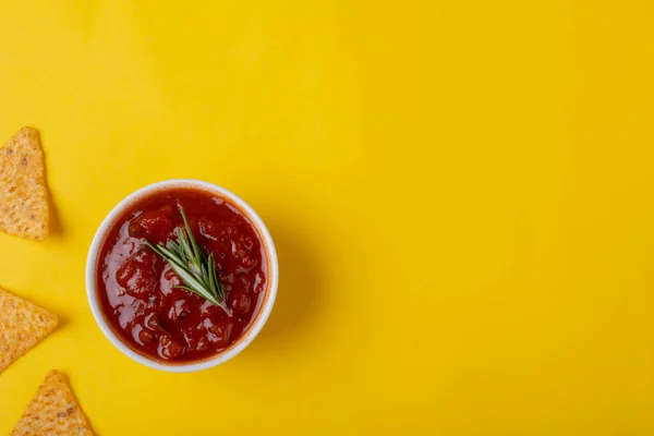 Red sauce with rosemary in bowl by nacho chips on yellow background with copy space. unaltered, unhealthy food, snack, dipping sauce, herb, crunchy and savory food.