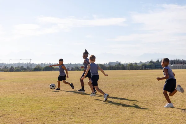 在阳光明媚的日子里 多种族小学生在学校足球场上与天空对抗踢足球 体育培训 团队体育和体育活动概念 — 图库照片