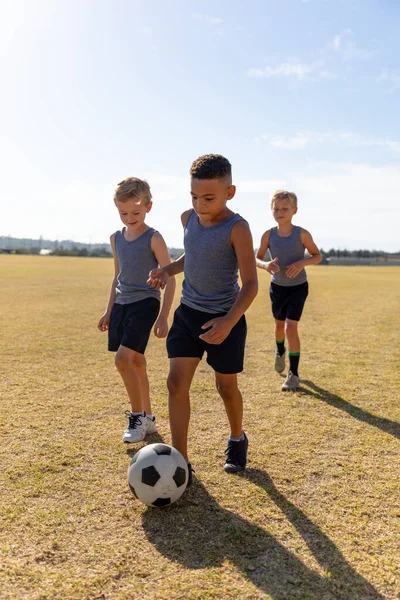 サッカー場で空を背景にサッカーをする多人種の小学生たち 変化のない子供時代教育スポーツトレーニングチームスポーツ活動のコンセプト — ストック写真
