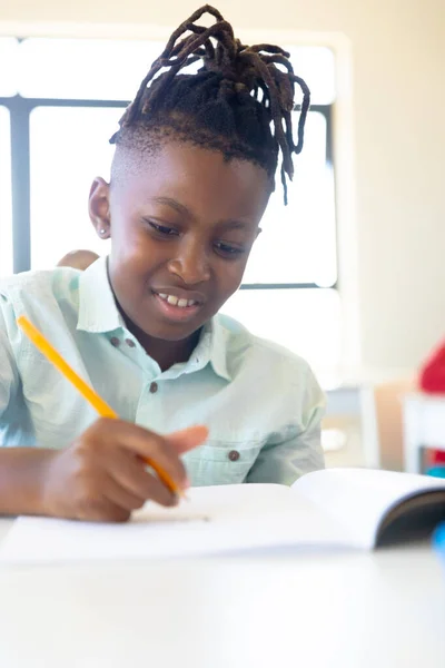 学校の机に座っている間に本に書いているアフリカ系アメリカ人の小学生を笑っています 子供時代教育学習文章を学校のコンセプトに戻し — ストック写真