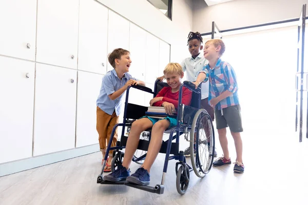 男子同級生が廊下に車椅子に腰掛けている姿は 元気な多人種間の男子学生 変化のない子供時代身体障害援助教育を学校のコンセプトに — ストック写真