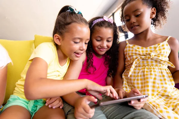 ソファに座っている間に 多人種の小学生の女の子がデジタルテーブルを使って笑っています 変化のない子供時代の無線技術一緒に教育を学校のコンセプトに戻し — ストック写真