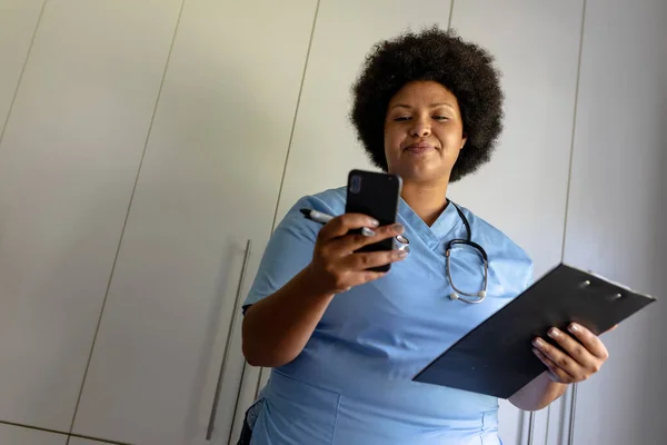 病院でクリップボードを保持しながら スマートフォンを使用してアフリカ系アメリカ人の中高年女性看護師 変更されず医療従事者無線技術専門知識職業概念 — ストック写真