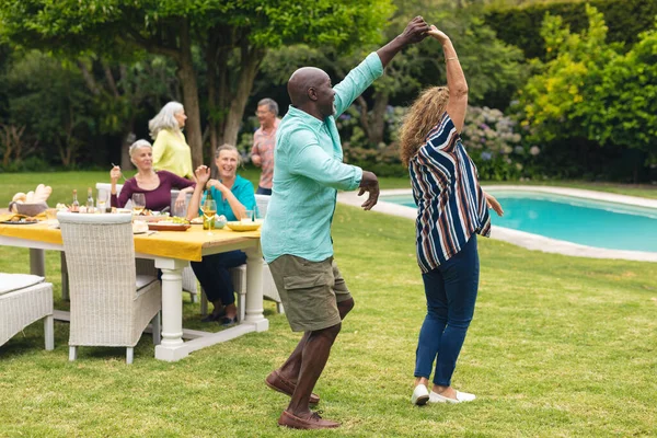 在后院里的聚会上 多种族的老夫妇一边跳舞 而朋友们则在餐桌边伴舞 生活方式 退休和后院派对 — 图库照片