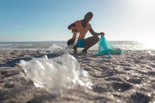African american man crouching while collecting garbage in plastic bag at beach on sunny day. unaltered, responsibility and environmental issues concept.
