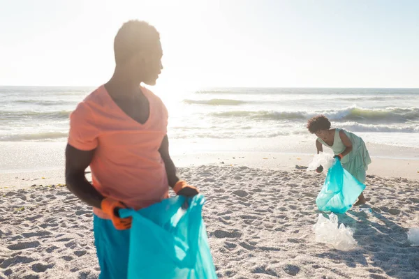ビーチからゴミを集めている彼女を見ながらビニール袋を持っているアフリカ系アメリカ人男性 変化のない連帯責任環境問題の概念 — ストック写真