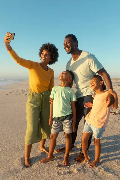 Happy african american woman taking selfie with family from smartphone at beach against blue sky. unaltered, family, lifestyle, togetherness, technology and holiday concept.