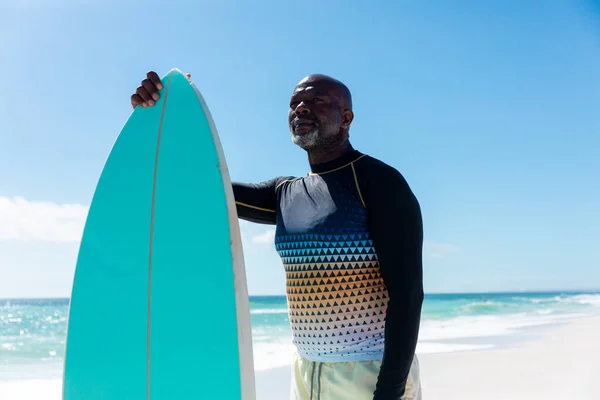 引退したアフリカ系アメリカ人の男性が青い空に対してビーチに立ってサーフボードを保持している 変化のないアクティブなライフスタイル水生スポーツ休日の概念 — ストック写真