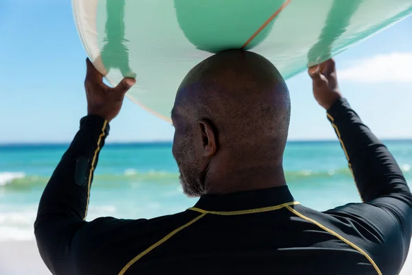 ビーチで頭の上にサーフボードを運ぶアフリカ系アメリカ人のはげ先輩の背面ビュー 変化のないアクティブなライフスタイル水生スポーツ休日の概念 — ストック写真