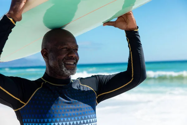 アフリカ系アメリカ人のはげ引退した老人がビーチでサーフボードを担いで微笑んだ 変化のないアクティブなライフスタイル水生スポーツ休日の概念 — ストック写真