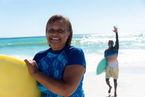 老人がビーチで手を振ってサーフボードを運ぶアフリカ系アメリカ人女性の笑顔の肖像画 変更されず団結し積極的なライフスタイル水生スポーツ休日の概念 — ストック写真