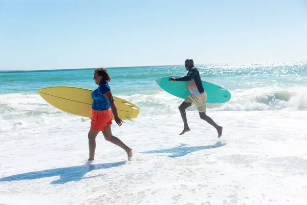 アフリカ系アメリカ人のカップルは コピースペースのある澄んだ空に対して海岸沿いのサーフボードで走っています 変更されず団結し積極的なライフスタイル水生スポーツ休日の概念 — ストック写真