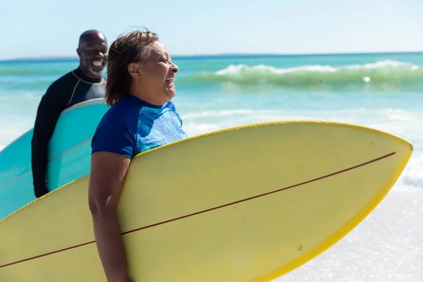 晴れた日にビーチを歩くサーフボードを運ぶ陽気なアフリカ系アメリカ人のシニアカップル 変更されず団結し積極的なライフスタイル水生スポーツ休日の概念 — ストック写真
