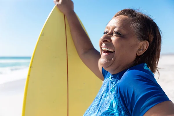 阳光灿烂的日子 快乐的非洲裔美国老年妇女站在沙滩上的黄色冲浪板旁 不变的 积极的生活方式 水上运动和假日观念 — 图库照片