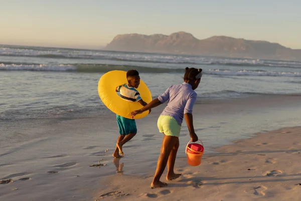 日没時にビーチで実行されているペアとインフレータブルリングと女の子と遊び心のあるアフリカ系アメリカ人の少年 変わらない 子供時代 ライフスタイル 楽しみ — ストック写真
