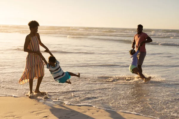 楽しいアフリカ系アメリカ人の若い両親は 日没の空に対して海岸で息子と娘を回転させます 変わらないビーチ 子供時代 ライフスタイル 楽しみと休日の概念 — ストック写真