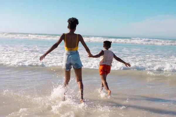 少女の手を取り 空に対して海で楽しむアフリカ系アメリカ人の若い母親の背面ビュー コピースペース 変更されていない ビーチ 子供時代 一緒に 楽しみ — ストック写真