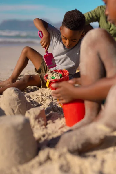 ビーチで父と妹と座っている間 アフリカ系アメリカ人の少年は砂 シャベルで遊んでいます 変化のないビーチ子供時代家族団結ライフスタイル楽しみ休日夏 — ストック写真