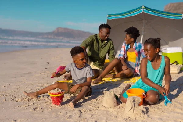 African american parents sitting under umbrella while children playing with sand, pails and shovels. summer, nature, unaltered, beach, childhood, family, togetherness, lifestyle, enjoyment, holiday.