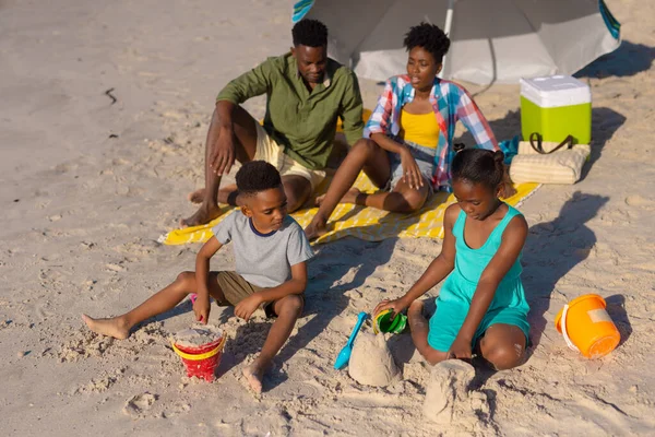 非洲裔美国父母坐在伞下 而孩子们在玩沙子时 他们的视角很高 生活方式 享受和假日 — 图库照片