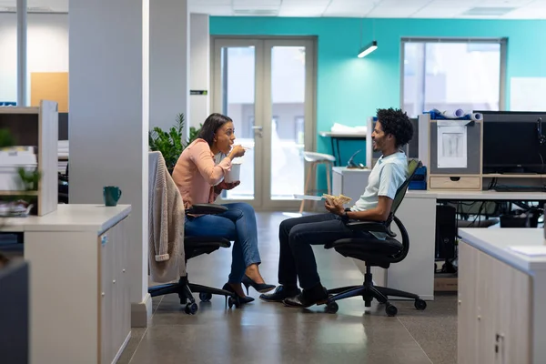 Multiracial professionals eating lunch together while sitting on chairs discussing at workplace. unaltered, business, food, teamwork and modern office concept.