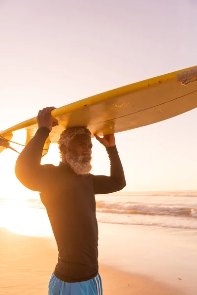 Afrykański Starszy Człowiek Niosący Deskę Surfingową Głowie Stojąc Naprzeciw Morza — Zdjęcie stockowe