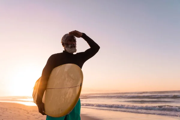 アフリカ系アメリカ人の老人は 澄んだ空に向かってビーチでサーフボードのシールドの目を保持ひげを生やした コピースペース 変更なし 水生スポーツ 休日とアクティブなライフスタイル — ストック写真