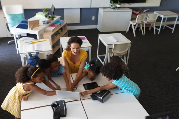 クラスでアフリカ系アメリカ人の小学生にラップトップを示す若い女性の白人教師 変更されていない 仮想現実シミュレータ 無線技術と学校の概念 — ストック写真