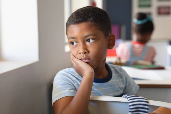 Biracial Základní Školák Rukou Bradě Dívá Pryč Zatímco Sedí Stolem — Stock fotografie