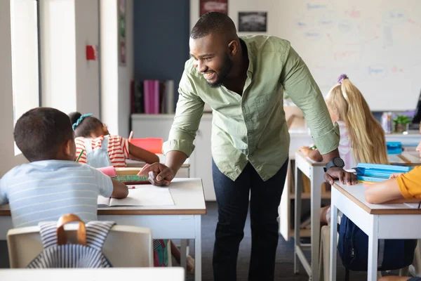 アフリカ系アメリカ人の若い男性教師がデスクでアフリカ系アメリカ人の小学生と話しているのを笑っている 変化のない教育学習子供時代教育職業学校の概念 — ストック写真