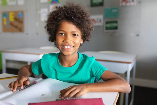 教室で机の上で勉強している巻き毛のアフリカ系アメリカ人の小学生の笑顔の肖像画 無修正 子供時代 学校のコンセプト — ストック写真
