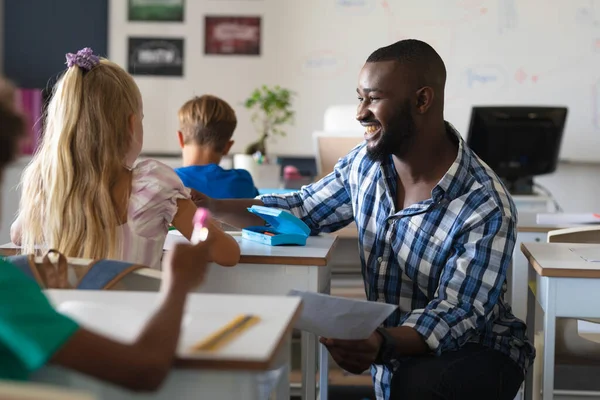 アフリカ系アメリカ人の若い男性教師が机の上で白人の小学生と話しているのを微笑んだ 変化のない教育コミュニケーション子供時代職業教育学校の概念 — ストック写真
