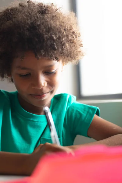 教室の机の上に座りながらペンを持っているアフリカ系アメリカ人の小学生の笑顔のクローズアップ 無修正 子供時代 学校のコンセプト — ストック写真