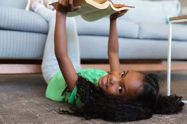 ソファの上に足で床に横たわっている間 アフリカ系アメリカ人の小学生の少女の本を読んでの肖像画 無修正 子供時代 リラックス 学校のコンセプト — ストック写真
