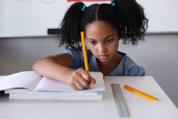 Biracial Elementary Schoolgirl Writing Book Desk While Studying Classroom Unaltered — Stock Photo, Image