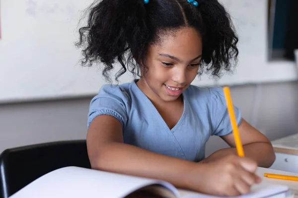 在课桌边读书的同时 微笑着小学女生在书上写字 集中精神和学校观念 — 图库照片