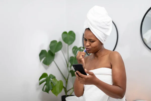 Африканська Середньостатистична Жінка Рушнику Використовуючи Смартфон Чистить Зуби Ванній Кімнаті — стокове фото