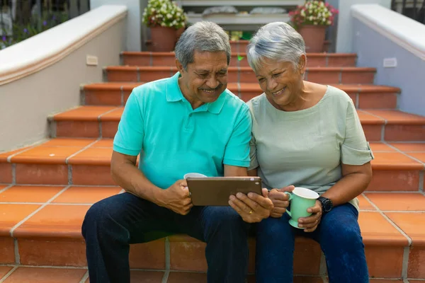 Smiling Biracial Senior Couple Holding Coffee Cups Using Digital Tablet —  Fotos de Stock