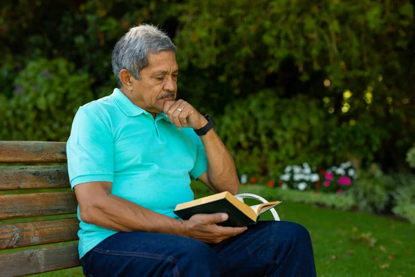 Biracial Senior Man Hand Chin Reading Book While Sitting Bench — Stockfoto