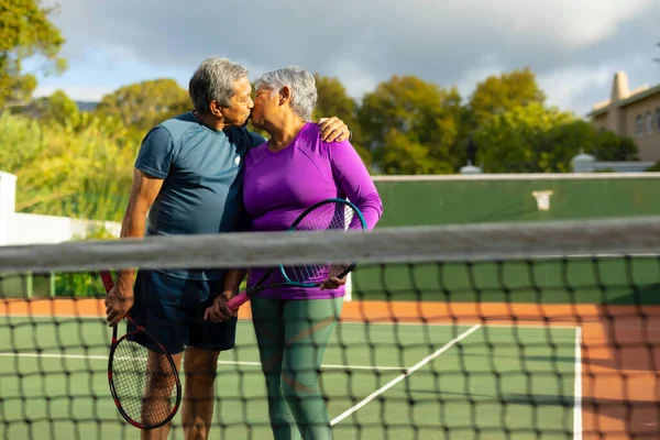 Biracial Loving Senior Couple Holding Rackets Kissing While Standing Tennis — Fotografia de Stock