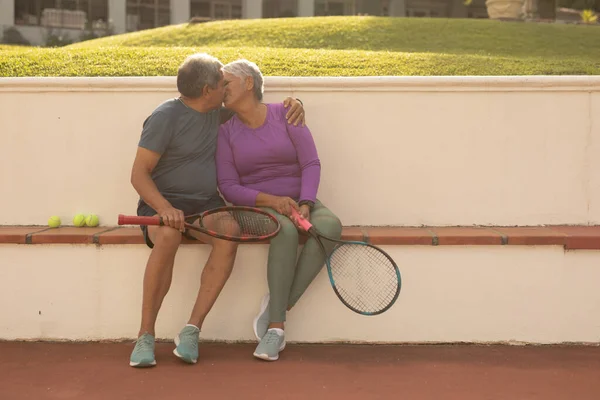 Romantic Biracial Senior Couple Holding Rackets Kissing While Siting Bench — Fotografia de Stock