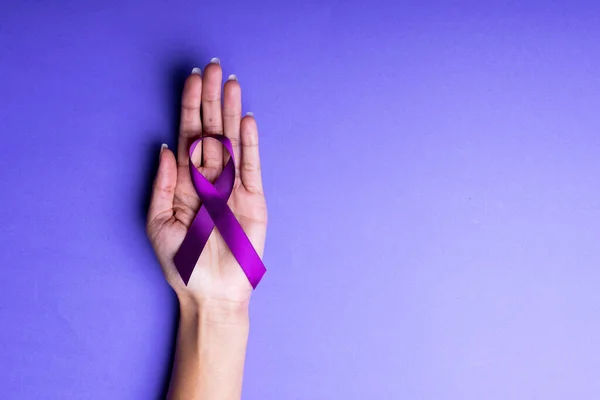 Cropped hand of woman holding purple awareness ribbon on blue background. copy space, epilepsy, pancreatic cancer, medical, awareness, support, healthcare and alertness.
