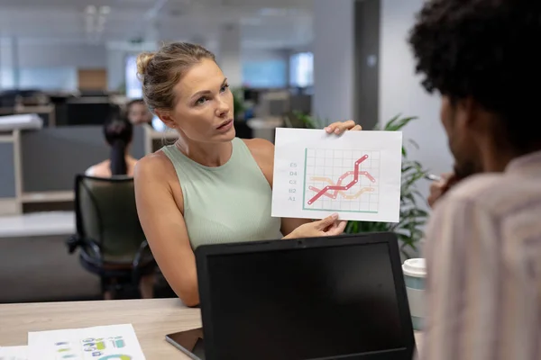 Confident caucasian businesswoman showing graph to biracial businessman during meeting in workplace. unaltered, business, teamwork, modern office and technology concept.
