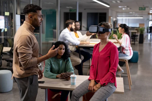 Hispanic businesswoman discussing with caucasian businesswoman wearing vr simulator in meeting. unaltered, business, teamwork, modern office and technology concept.