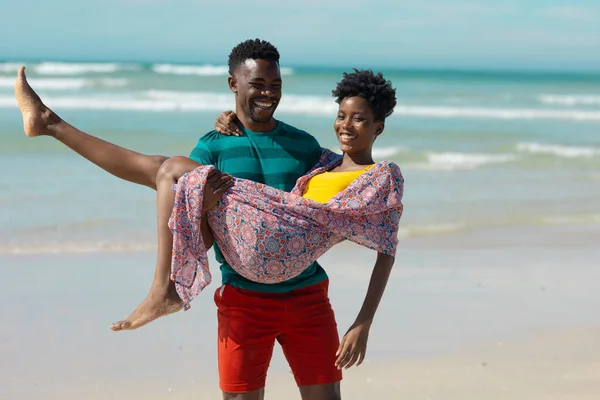 Happy african american young man carrying young girlfriend while standing at beach against sea. summer, nature, unaltered, beach, love, togetherness, lifestyle, enjoyment and holiday concept.