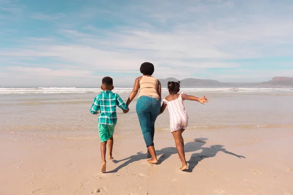 非洲裔美国祖母牵着男孩和女孩的手在海滩上迎着天空奔跑的后视镜 假日观念 — 图库照片