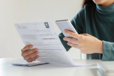 Asyalı müşteri kadın, internet üzerinden tarama ve ödeme için cep telefonu kullanıyor. Aile bütçesi faturası ev ofisinin üzerinde, para tasarrufu, yatırım, işletme finansmanı, masraf konsepti.