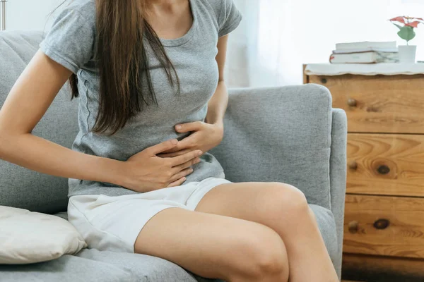 asian young woman suffering stomach ache sitting on couch in living room at home, people painful stomachache, gynecology, menstrual pain , medical and health care concept