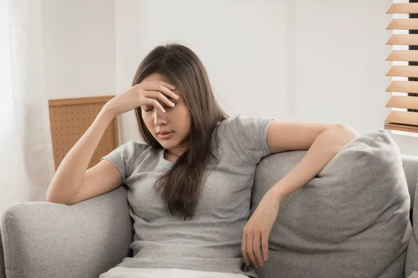young asian woman touching forehead having headache migraine and upset with problem feel stressed sitting on couch in living room at home, people painful, depression, medical and health care concept